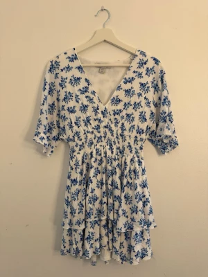 Vit kortklänning med blå blommor - Söt vit kortklänning med blått blommigt mönster och v-ringning. Klänningen har volangdetaljer i kjolen, resår i midjan och korta ärmar. Perfekt för varma dagar och somriga tillfällen.