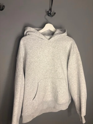 Ljusgrå basic hoodie med huva - En stilren ljusgrå hoodie med huva och stor magficka. Tröjan har en avslappnad passform, ribbade muddar vid ärmslut och nederkant samt klassisk känguruficka framtill. Perfekt för en chill och enkel look.
