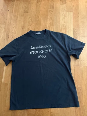Svart Acne Studios t-shirt - Säljer en svart t-shirt från Acne Studios med tryck i vitt framtill där det står 'Acne Studios STOCKHOLM 1996'. Klassisk passform och rund halsringning. Perfekt för dig som gillar stilrena och ikoniska plagg.