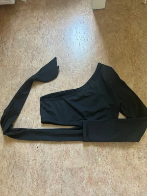 Svart oneshoulder topp med knytband - Trendig svart oneshoulder topp med lång ärm och ett långt knytband som ger en unik look. Perfekt för dig som vill sticka ut med en cool och modern stil. Tillverkad i mjukt material som sitter snyggt på kroppen.