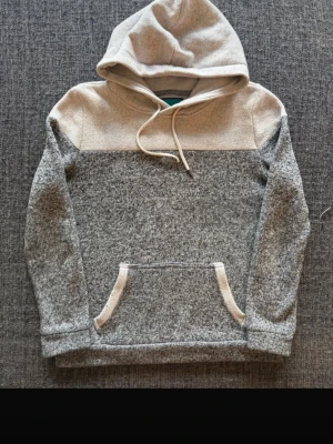  hoodie med huva - Säljer en stilren gråmelerad hoodie med ljusgrå partier upptill och på fickkanterna. Hoodien har en stor huva med dragsko och en klassisk magficka. Materialet ät mjukt och mysigt, perfekt för chill dagar.