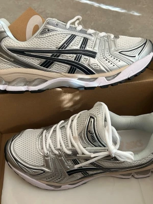 Vita och silver Asics sneakers - Snygga sneakers från Asics i vitt och silver med svarta detaljer. Skorna har en sportig design med mesh- och syntetmaterial, samt klassiska Asics-loggor på sidorna. Perfekta för en trendig och bekväm stil. Tjock vit sula och vita skosnören.