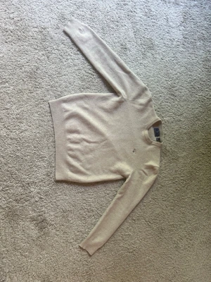 Beige stickad tröja med rund hals - Säljer en stilren beige stickad tröja i lamm ull med rund hals och långa ärmar. Tröjan har en diskret broderad detalj på bröstet och ribbade muddar vid ärmslut och nederkant. Perfekt för lager på lager och enkel att matcha.