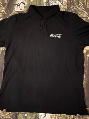 Svart pikétröja med Coca-Cola tryck  - Snygg svart pikétröja med klassisk krage och korta ärmar. Storlek XL men är egentligen mindre. Passar dig som också bör storlek M och L. På bröstet finns ett vitt Coca-Cola tryck som ger en retro känsla. Perfekt till jeans eller shorts för en avslappnad look.