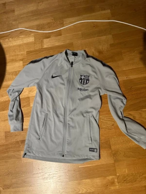 Grå Nike FC Barcelona träningsjacka - Snygg grå träningsjacka från Nike med FC Barcelona-logga och Rakuten-tryck på bröstet. Jackan har hel dragkedja, ståkrage och svarta detaljer på axlarna. Perfekt för fotbollsträning eller som sportig vardagsstil.