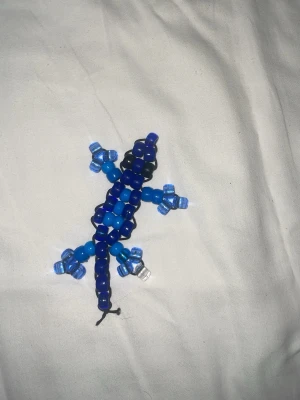 KANDI Blå ödla  - Säljer en handgjord ödla i olika blå nyanser av plastpärlor. Ödlan har detaljer i ljusblått och genomskinligt, och är trädd på svart tråd. Perfekt som dekoration till väska, nyckelring eller smycke.