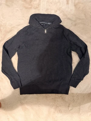 Mörkgrå tröja med halv zip-up - Mörkgrå tröja med hög krage och halv dragkedja framtill. Tröjan har långa ärmar och ribbade muddar vid ärmslut och nederkant. Perfekt för en avslappnad och stilren look.