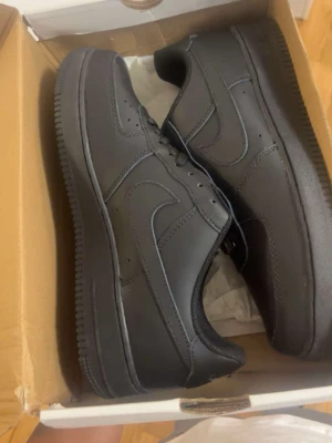 Nike Air Force 1 svarta sneakers - Nike Air Force 1 sneakers i helsvart läder med klassisk låg siluett. Skorna har perforerad tå, svart sula och matchande svarta skosnören. Perfekta för en clean och stilren look.