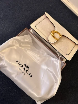 Vit kuvertväska från Coach - Snygg vit kuvertväska från Coach i strukturerat läder med guldfärgat C-spänne framtill. Väskan har svarta kanter och klassisk, stilren design. Levereras i originalbox och med dustbag från Coach.