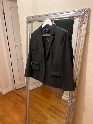 Mörkgrå kavaj från Stockh - Snygg mörkgrå kavaj från Stockh med klassisk krage och två fickor med lock framtill. Kavajen har en enkelknäppning och är helfodrad med diskret mönstrat innerfoder. Perfekt för en stilren och modern look.