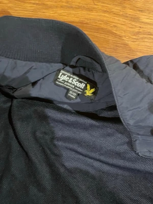 Mörkblå vindjacka från Lyle & Scott - Snygg mörkblå vindjacka från Lyle & Scott med dragkedja, ståkrage och två fickor framtill. Jackan har det klassiska gula logobroderiet på bröstet och elastiska muddar vid ärmslut och midja. Perfekt för blåsiga dagar och enkel att matcha. Passar med alla stilar. Funkar både vår och höst. Skriv för att diskutera pris