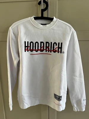 Vit sweatshirt från Hoodrich - Vit sweatshirt från Hoodrich med svart och rött tryck på både fram- och baksidan. Tröjan har rund halsringning, långa ärmar och ribbade muddar. Detaljer som patch nertill och broderade texter på ärmen och nederkanten ger en streetwear-känsla.