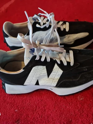 Svarta och vita New Balance sneakers - Säljer ett par snygga New Balance sneakers i svart och vitt. Skorna har en stor vit N-logga på sidan, vita skosnören och en vit sula. Ovandelen är i mocka och textil, vilket ger en sportig och modern look. Perfekta för dig som gillar streetwear och vill sticka ut.