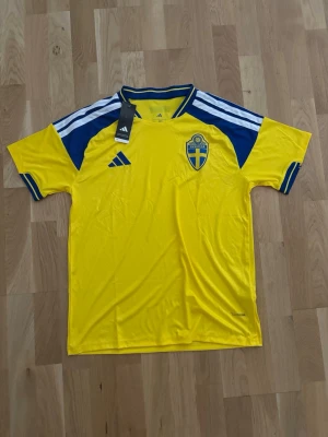 Svensk landslagströja Adidas gul/blå - Gul och blå svensk landslagströja från Adidas med klassiska tre ränder på axlarna och Sverigelogga på bröstet. Tillverkad i lätt och ventilerande material, perfekt för fotboll eller träning. Rund halsringning med blå detaljer.