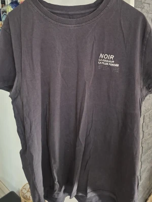 Svart t-shirt med tryck - Svart t-shirt i bomull med texttryck på bröstet där det står 'NOIR LA COULEUR LA PLUS FONCÉE'. Klassisk rund halsringning och korta ärmar. Perfekt till jeans eller shorts för en enkel och stilren look.