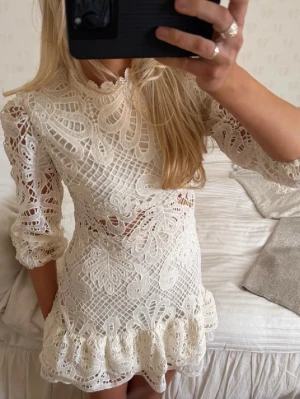 Vit spetsklänning med volang - Superfin vit kortklänning i spets med hög hals och trekvartsärm. Klänningen har vackra broderade detaljer, volang nedtill och en figurnära passform. Perfekt för sommarens alla tillfällen!