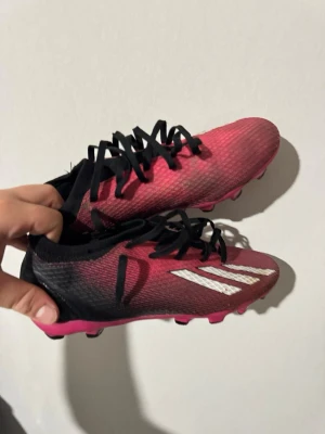 Rosa och svarta adidas x speedportal ag - Snygga fotbollsskor i rosa och svart med vita detaljer och svarta snören. Skorna har en låg profil och är tillverkade i ett lätt syntetmaterial för bra bollkontroll. Dobbarna på sulan ger grymt grepp på gräsplanen. Perfekta för dig som vill sticka ut på planen!