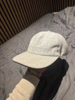 Vit keps från Fendi med mönster - Snygg vit keps från Fendi med diskret mönster i tyget och böjd skärm. Kepsen har justerbar rem baktill och är tillverkad i ett tygmaterial som känns lyxigt. Perfekt för att lyfta din outfit med en exklusiv touch.