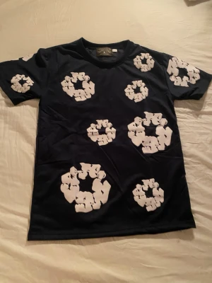 DENIM TEARS set - Cool svart t-shirt med stora vita blommönster över hela plagget. T-shirten har klassisk passform och korta ärmar. Perfekt för dig som vill sticka ut med ett unikt mönster och enkel stil.