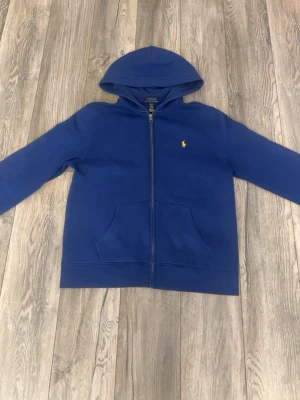 Blå hoodie från Polo Ralph Lauren - Snygg blå hoodie från Polo Ralph Lauren med dragkedja framtill och klassisk huva. Tröjan har fickor på magen och den ikoniska lilla gula loggan broderad på bröstet. Tillverkad i mjukt material som känns skönt mot huden. Perfekt för en avslappnad och stilren look.
