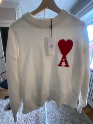 Vit stickad tröja från Ami Paris - Vit stickad tröja från Ami Paris med röd hjärtlogga och bokstaven A på bröstet. Tröjan har ribbstickad krage, ärmslut och nederkant. Oversized passform och mjukt material som känns lyxigt och trendigt.