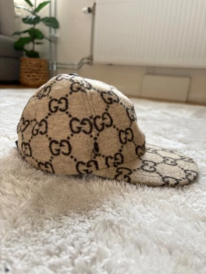 Fluffig gucci keps - Säljer en beige keps från Gucci med det klassiska GG-monogrammet i svart över hela kepsen. Kepsen har böjd skärm och en knapp på toppen. Materialet är en ullblandning som ger en lyxig känsla och unik look.