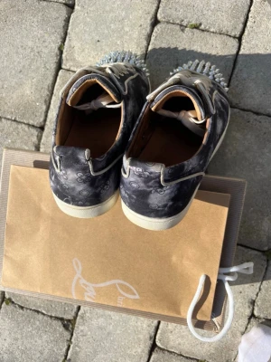 Christian Louboutin  - Unika Christian Louboutins med vitt mönster och silverfärgade nitar på tån. Skorna har en cool röd sula, kontrasterande design. Perfekta för dig som vill sticka ut och gillar detaljer som drar blickarna till sig.