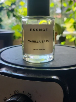 ESSNCE Vanilla Sass' Eau de Parfum💕 - Fräsch parfym från ESSNCE med doften Vanilla Sass'. Kommer i en genomskinlig, rund glasflaska med svart lock och enkel etikett. Innehållet är ljust gult och flaskan har en modern, minimalistisk design som passar perfekt på parfymhyllan.