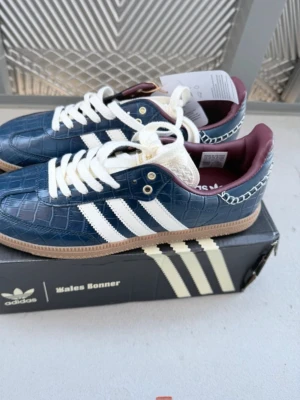 Adidas Wales Bonner blå sneakers - Unika sneakers från Adidas x Wales Bonner i blått med krokodilpräglat läder, vita ränder och detaljer samt vinröda insidor. Skorna har vita snören, beige sula och gulddetaljer vid snörningen. Perfekt för dig som vill sticka ut med stilrena och trendiga sneakers.