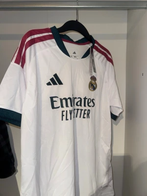Real Madrid matchtröja Adidas - Säljer en vit Real Madrid matchtröja från Adidas med mörkgröna detaljer och vinröda ränder på axlarna. Tröjan har klubbmärke och Adidas-logga på bröstet samt 'Emirates Fly Better' tryckt framtill. 