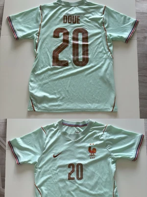 Frankrike bortaställ Nike #20 Doue - Ljus turkos fotbollströja från Nike med Frankrikes landslagsemblem och nummer 20 på både fram- och baksida. Namnet 'DOUE' står på ryggen. Tröjan har korta ärmar med randiga muddar och tunna röda detaljer längs sidorna..