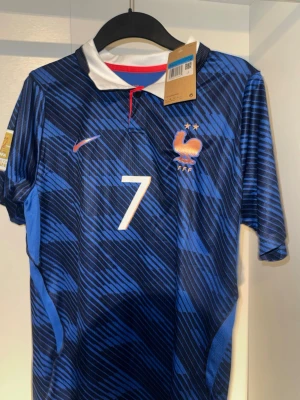 Frankrike fotbollströja Dembele #7 - Säljer en blå fotbollströja från Frankrikes landslag med mönstrad design, vita detaljer och nummer 7 på bröstet och ryggen. På ryggen står det 'Dembele'. Tröjan har korta ärmar, vit krage och broderad tupp på bröstet.