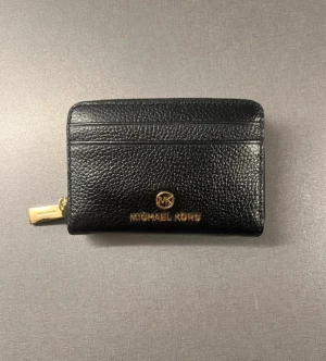 Svart Michael Kors plånbok  - Snygg svart plånbok från Michael Kors, guldfärgade detaljer och logotyp framtill. Plånboken har flera kortfack och dragkedja i sidan. Perfekt storlek för att ha med sig i fickan eller väskan.