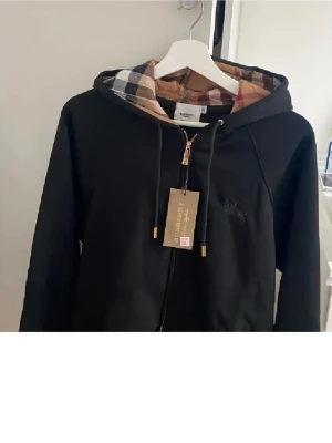 Svart hoodie med rutigt foder - Snygg svart hoodie med dragkedja och huva. Insidan av huvan har ett klassiskt beige, svart och rött rutmönster. Guldiga detaljer på dragkedjan och snörändarna ger en exklusiv känsla. Perfekt för dig som vill ha en stilren men ändå unik hoodie.