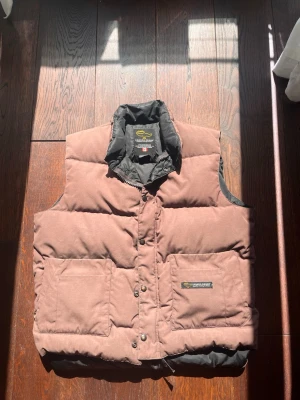 Brun dunväst från Canada Goose - Säljer en brun dunväst från Canada Goose med hög krage och stora framfickor. Västen är quiltad och har tryckknappar framtill. Perfekt för lager-på-lager under kyliga dagar. Klassisk och stilren design med svart insida.
