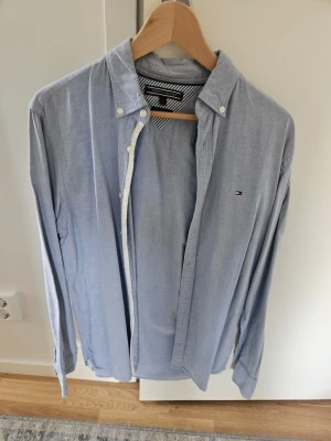 Ljusblå skjorta från Tommy Hilfiger - Klassisk ljusblå skjorta från Tommy Hilfiger med knappkrage och diskret logga broderad på bröstet. Skjortan har långa ärmar och knappar framtill. Perfekt till jeans eller chinos för en clean look.