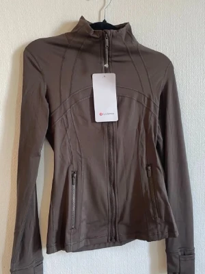 Brun träningsjacka från Lululemon - Snygg brun träningsjacka från Lululemon med hög krage och hel dragkedja framtill. Jackan har två fickor med dragkedja och dekorativa sömmar som ger en sportig look. Tillverkad i ett stretchigt och mjukt material som passar perfekt till träning eller promenader.