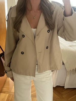 Kort beige trenchcoat med knappar - Stilren kort trenchcoat i beige med dubbelknäppning och stora svarta knappar. Finns tillhörande band till. Är strl L men passar även mig som brukar ha S.