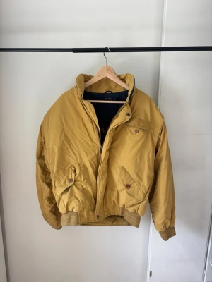 Gant vintage vindjacka - Snygg gul Gant vindjacka i retrostil med hög krage och dragkedja framtill. Jackan har två stora fickor med lock och knapp, samt ribbade muddar vid ärmslut och midja. Materialet är glansigt och jackan har en avslappnad passform.
