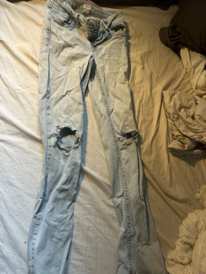 Ljusblå slitna jeans med brodyr -  Nelly jeans Säljer ett par ljusblå jeans med slitningar över knäna och broderade detaljer i rött på bakfickorna. Jeansen har klassisk femficksmodell bootcut och är tillverkade i denim. Perfekta för en avslappnad och trendig look. En defekt på sista bilden
