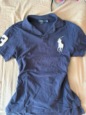 Marinblå pikétröja från Polo Ralph Lauren - Klassisk marinblå pikétröja från Polo Ralph Lauren med vit broderad logga på bröstet och siffran 3 på ärmen. Tröjan har krage, korta ärmar och knäppning vid halsen. Perfekt till en avslappnad men ändå stilsäker look.