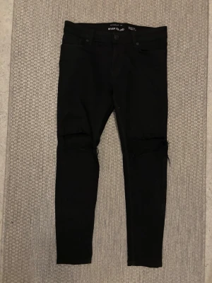 Svarta skinny jeans River Island - Säljer ett par svarta skinny jeans från River Island med slitningar vid knäna. Jeansen har fem fickor och hög midja. Snygg passform och stretchigt material som sitter tajt hela vägen ner.