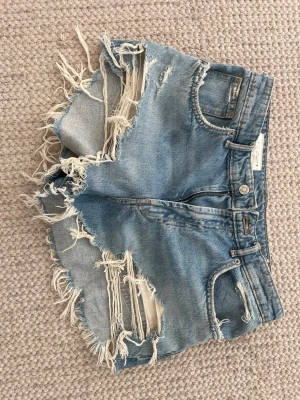 Blå slitna jeansshorts med fransar - Säljer ett par blå jeansshorts med råa kanter och stora slitningar framtill. Shortsen har fem fickor, dragkedja och knapp i midjan. Perfekta för sommaren och ger en avslappnad, trendig look.