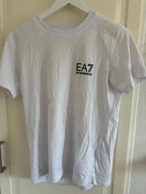 Vit EA7 t-shirt i bomull - Snygg vit t-shirt från EA7 Emporio Armani med svart logga på bröstet. Klassisk rund halsringning och korta ärmar. Tillverkad i mjuk bomull som känns skön mot huden. Perfekt till jeans eller shorts för en clean och stilren look.