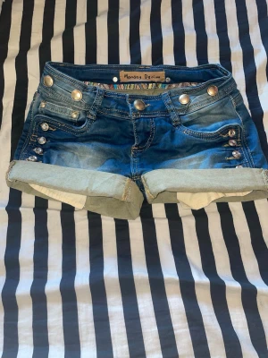 Blå denimshorts från Monday Denim - Snygga blå denimshorts från Monday Denim med uppvikta benslut och dekorativa knappar på sidorna. Shortsen har klassiska fickor fram och bak samt detaljerade sömmar och nitar. Perfekta för varma dagar och enkel att matcha med olika toppar.