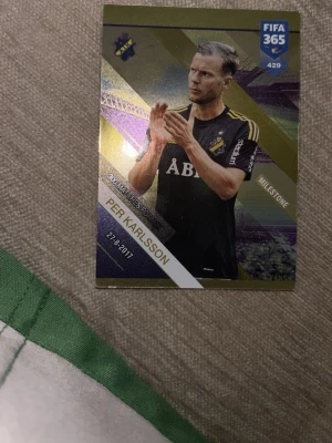 FIFA 365 Adrenalyn XL Per Karlsson kort - Samlarkort från FIFA 365 Adrenalyn XL med Per Karlsson i AIK-tröja. Kortet har guldfärgade detaljer, svart och lila bakgrund samt text om milstolpe. Baksidan har spelplan, färgade staplar och en aktiveringskod. Tillverkat av Panini.