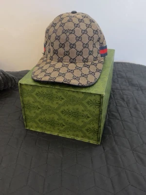 Beige keps med GG-mönster från Gucci - Säljer en beige keps från Gucci med det klassiska GG-monogrammet över hela kepsen. Kepsen har böjd skärm och en röd-grön rand på sidan. Materialet är canvasliknande och kepsen har en stilren och exklusiv look. Pris kan diskuteras!