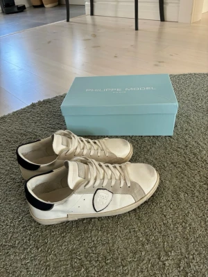 Vita Philippe Model sneakers låga - Snygga vita sneakers från Philippe Model med beige mockadetaljer och svart häl. Klassisk låg modell med vita skosnören och logotyp på sidan. Ovandelen är i läder och mocka, sulan är vit med en något smutsad look för en trendig vibe.
