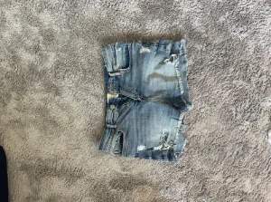 Blå slitna jeansshorts med fransar - Säljer ett par blå jeansshorts med slitna detaljer och fransig kant. Shortsen har klassisk femficksdesign och dragkedja framtill. Perfekta för sommaren och ger en avslappnad vibe.