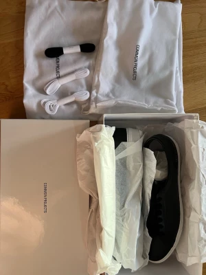 Svarta lädersneakers Common Projects - Stilrena svarta sneakers i läder från Common Projects med vit sula och klassisk låg profil. Skorna har matchande svarta skosnören och diskret guldprägling på hälen. Medföljer extra vita skosnören och dustbags.